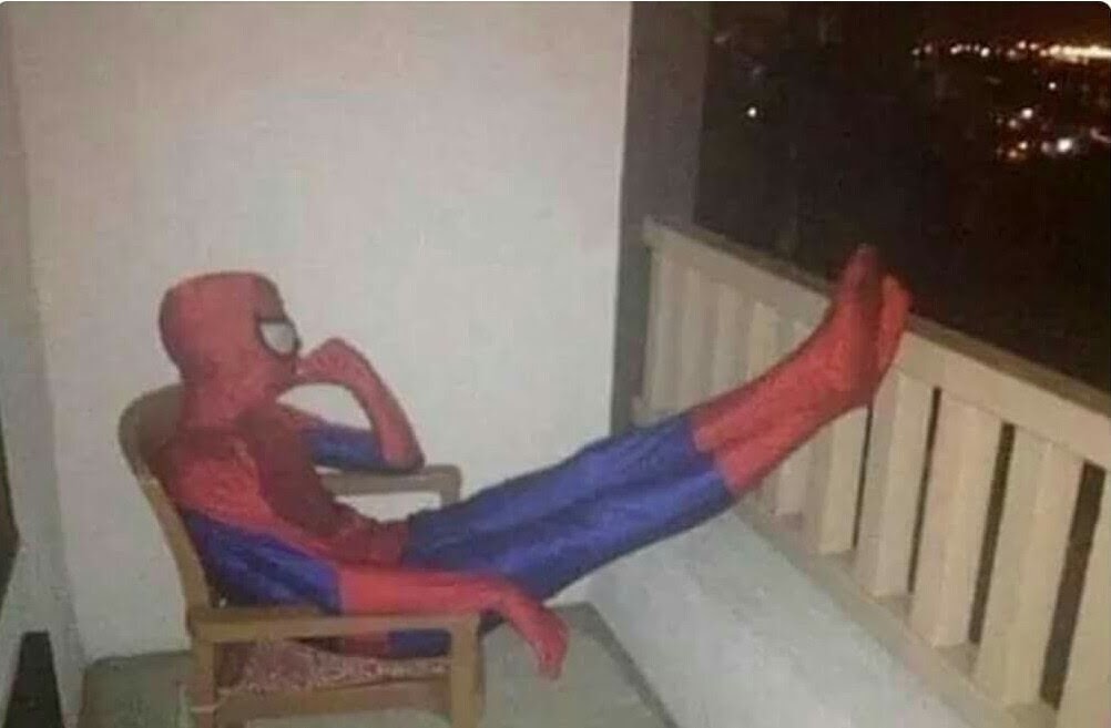 homem-aranha sentado na cadeira, com as pernas esticadas e apoiadas na janela com a mão no queixo pensativo